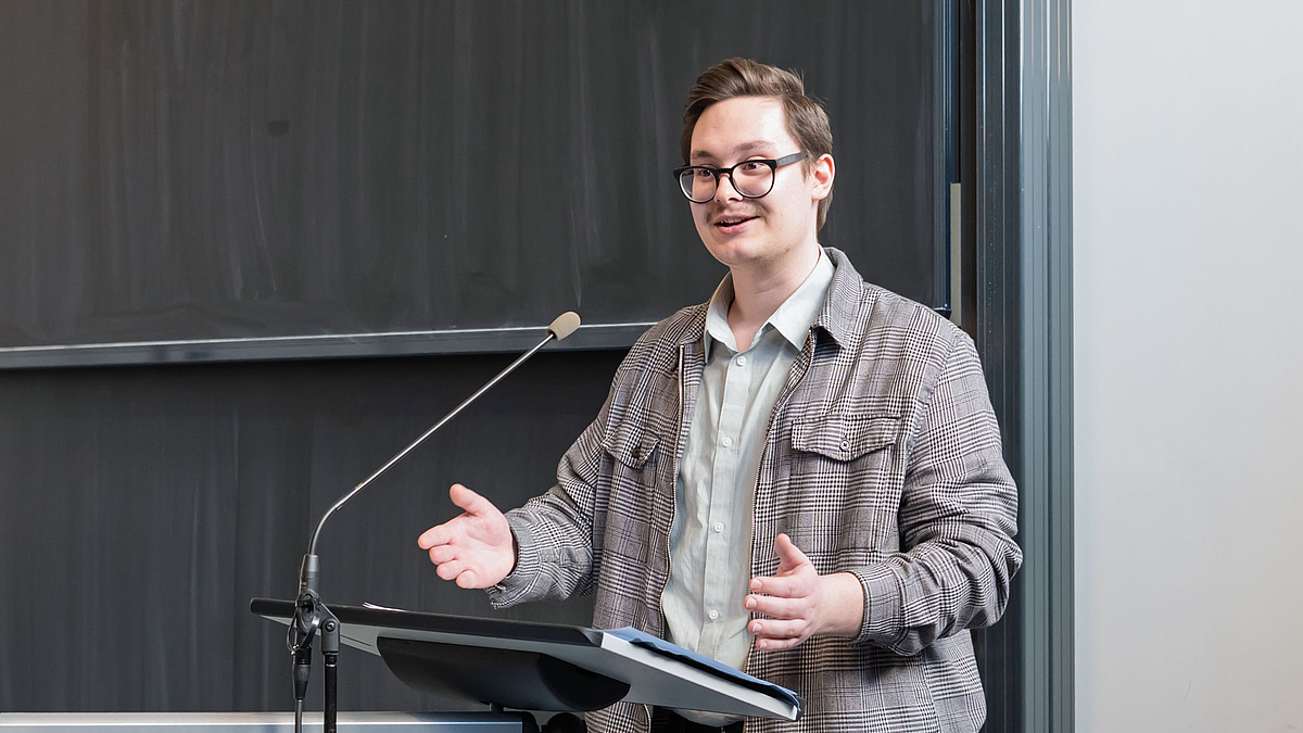 Leon Matschkewitz vom Studierendenrat stimmt seine neuen Kommilitonen ein und wirbt für das Engagement in den studentischen Gremien: „Hier zu studieren ist wie eine große Familienfeier. " Eine jüngere männliche Person am Rednerpult vor einer Tafel.