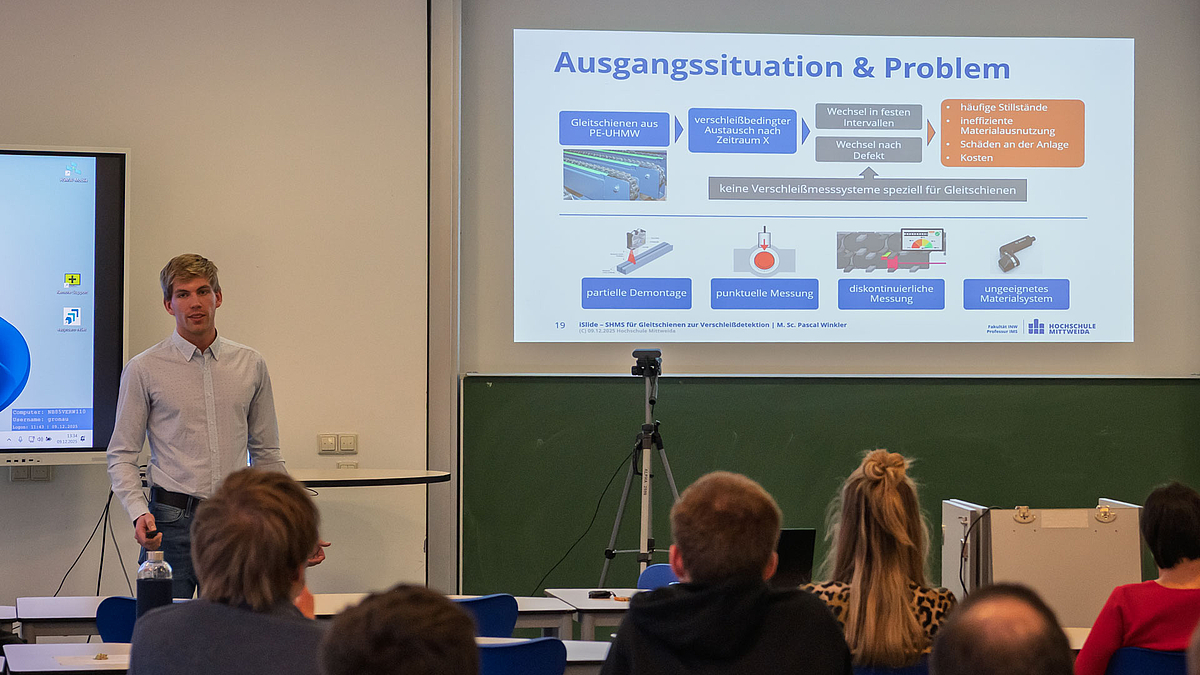 Platz drei: Jim Köcher präsentiert für seinen Kollegen Pascal Winkler. Männliche Person vor projizierter Powerpointpräsentation. Im Vordergrund Köpfe des Publikums von hinten.