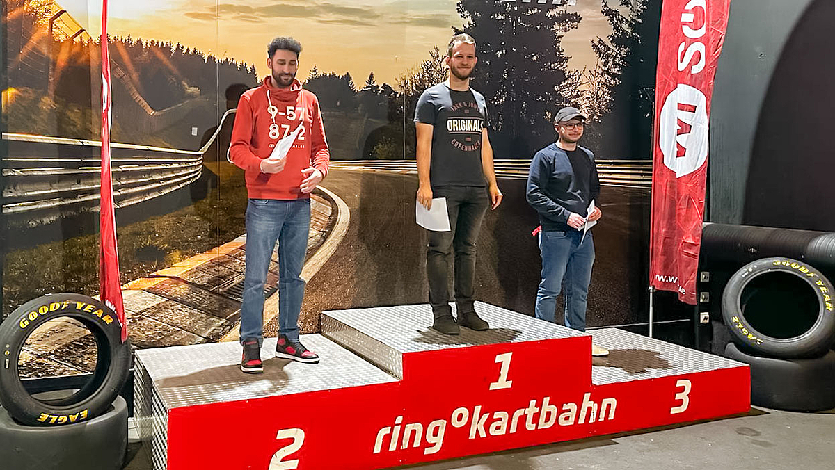 Drei junge männliche Personen stehen auf einem dreistufigen Siegerpodest und halten Urkunden in den Händen. Auf dem Podest ist weiß auf rot „Ring Kartbahn“ zu lesen. Rennreifen sind links und rechts vom Podest drapiert. Der Hintergrund ist die Abbildung eines Ausschnitts einer Rennkurve mit Leitplanken.