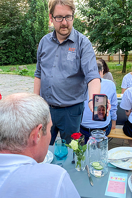 Auf einer Terrasse vor parkähnlichen Hintergrund stehen Biertischgarnituren. An ihnen sitzen Personen, die nur von hinten zu sehen sind.  Eine männliche Person hält stehend ein Smartphone in Richtung einer weiteren männlichen Person am Tisch im Vordergrund. Auf dem Bildschirm des Smartphones ist eine dritte männliche Person zu sehen.