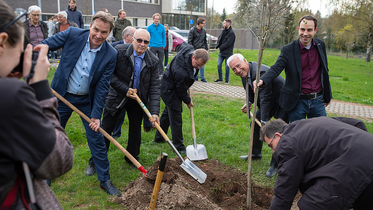 Die Gruppe der Ehrengäste der Gebäudebenennung zum "Horst-Exner-Bau" steht mit Spaten in den Händen vor einem Erdloch. Ein Mann im Anzug hält den Baum, dessen Wurzeln mit Erde bedeckt werden, aufrecht.