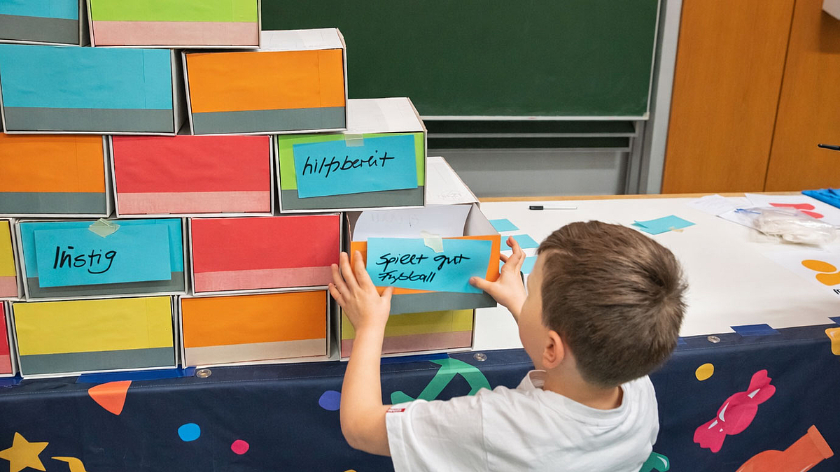 Ein Kind steht vor einem Tisch mit gestapelten, bunten Schubladen. Die Schubladen sind mit verschiedenen Eigenschaften beschriftet, darunter 'hilfsbereit' und 'lustig'.