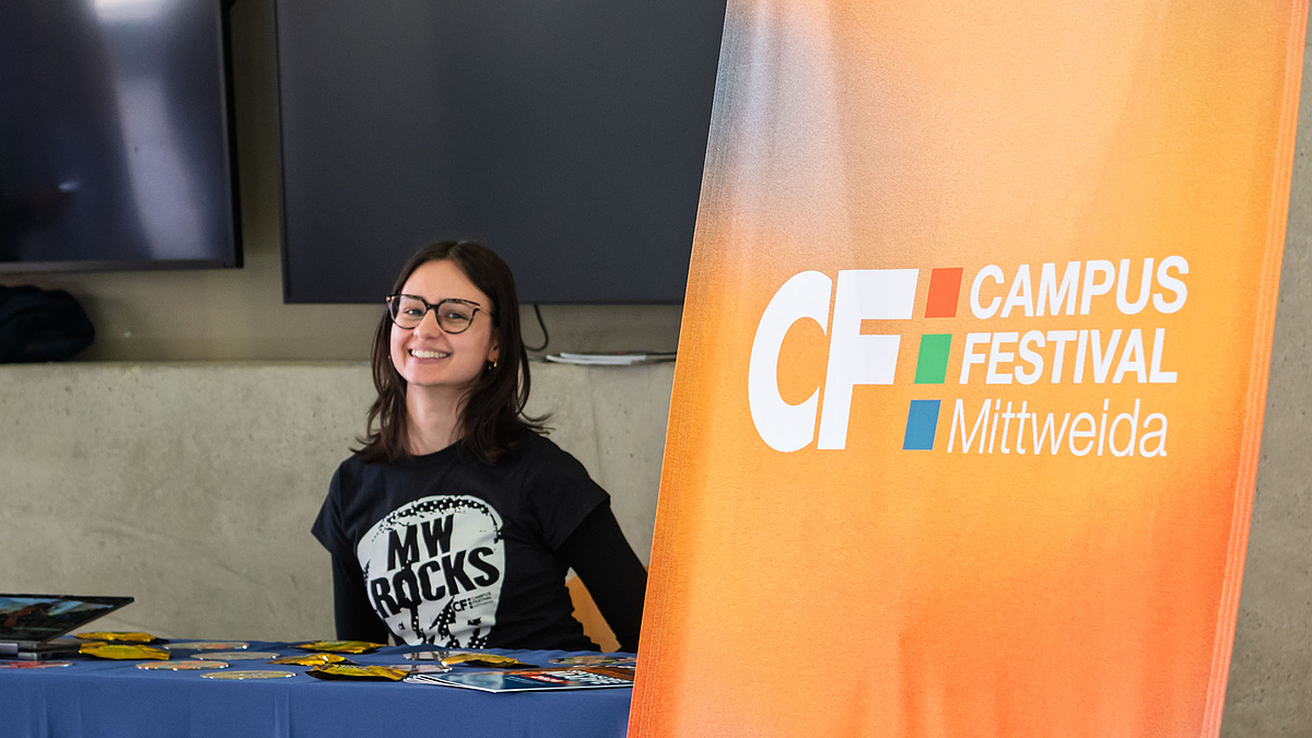 Eine junge weibliche Person mit "MW rocks"-Polo sitzt fröhlich lächeld an einem Tisch. Neben ihr im Bildvordergrund setht ein "Campusfesitival Mittweida"-Rollup