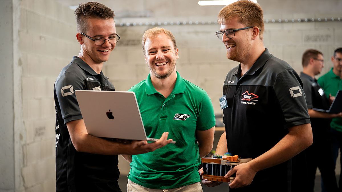 Drei männliche Personen schauen lachend auf einen geöffeneten Laptop