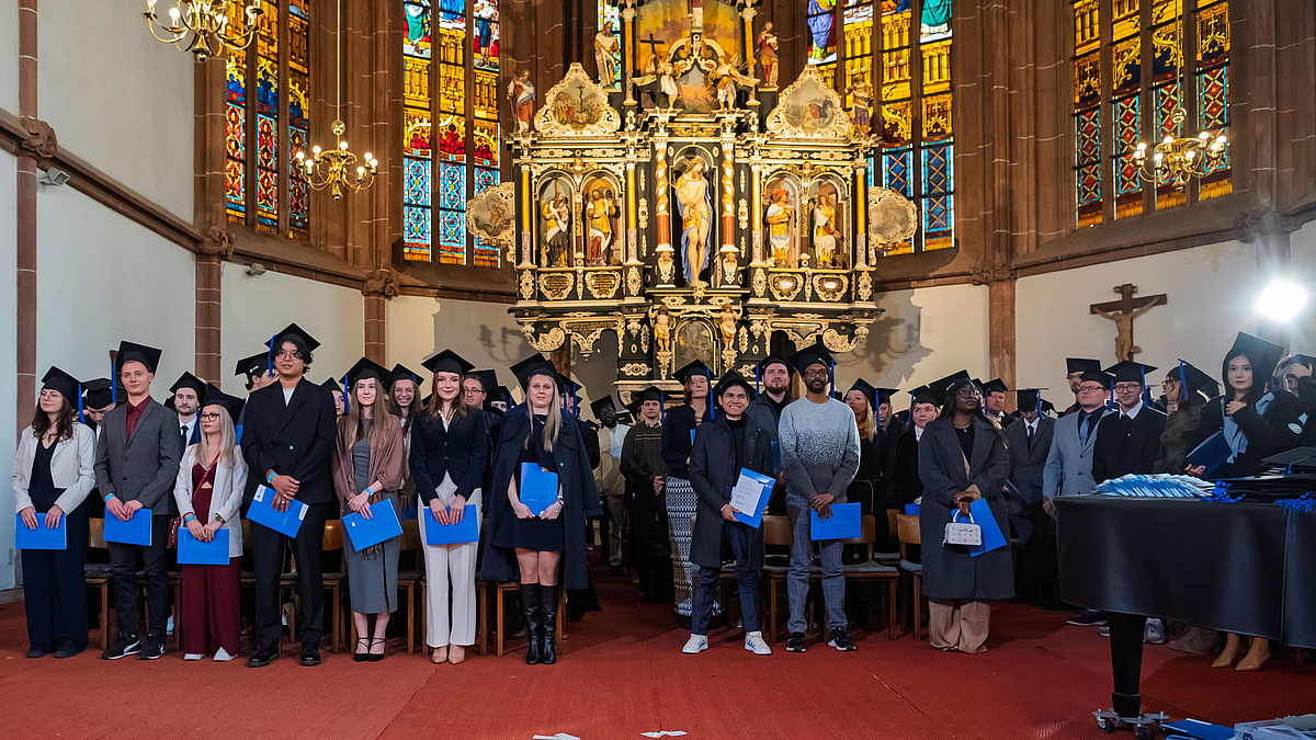 Absolvent:innen mit Hüten und blauen Urkundenmappen stehen im Altarraum.