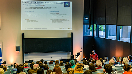 Wissenschaftler wie Professor Thomas Villmann kennen ChatGPT schon länger. Der seit Dezember 2022 für jedermann zugängliche Chatbot ist aber sicher ein Grund für das große Interesse auch von Nichtwissenschaftler:innen . Blick von hinten in den Hörsaal über die Köpfe des Publikums auf den männlichen Dozenten. Schräg über ihm ist eine Folie projiziert mit der Überschrift: „Anwendungen von KI und maschinellem Lernen – III – ChatGPT & Co“ und „Was ist, was kann ChatGPT ¬ nicht?“