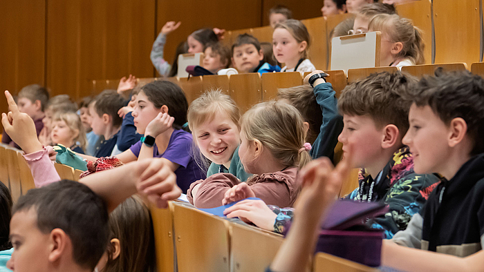Gruppe von Kindern in den ansteigenden Bankreihen des Hörsaals. Einige melden sich mit gestreckten Armen, während andere miteinander sprechen und lachen.