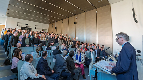 Ein Hörsaal voller frisch immatrikulierter Erstsemester an der Hochschule Mittweida. Im akademischen Alltag ist es meistens umgekehrt: Die Dozent:innen stehen und die Studierenden sitzen. Stehende junge Personen in den ansteigenden Bankreihen eines Hörsaals. In den unteren Reihen sitzen ältere Personen, die sich zu ihnen umdrehen.