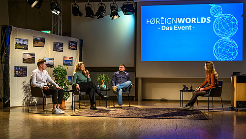 Nach dem Auslandssemester: Paul, Gina Louise und Max (v.l.) berichten Moderatorin Jule im FOREIGNWORLDS-Talk aus Spanien, Finnland und Rumänien. Das Foto zeigt auf der linken Seite drei junge Menschen auf Sesseln vor einer Wand mit Reisebildern sitzend. Die junge Frau in der Mitte spricht gerade in ihr Mikrofon. Rechts im Bild ist die Moderatorin vor einer großen Leinwand zu sehen, auf der neben drei stilisierten transparenten Weltkugeln vor blauem Hintergrund die Worte „FOREIGNWORLDS – Das Event“ zu lesen sind.