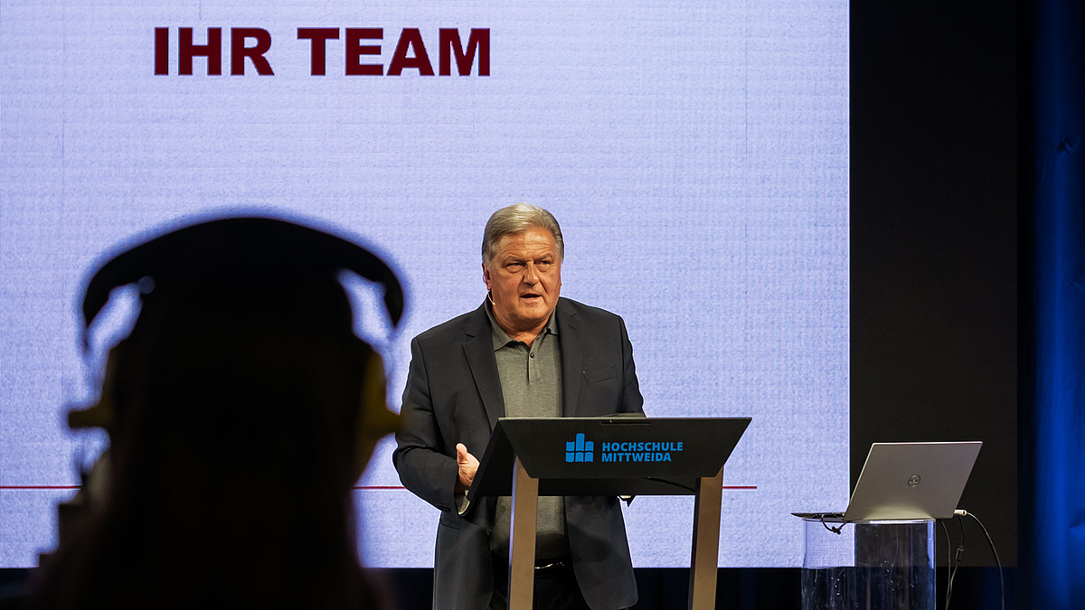 Blick an einer Studiokamera im Schatten vorbei auf den Referenten. Auf dem Screen hinter ihm ist "Ihr Team" zu lesen.