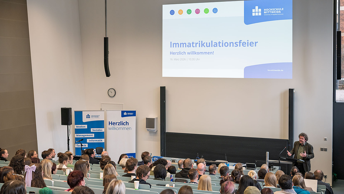 Blick vom oberen Ende des Hörsaals über die Köpfe der Zuhörenden hinweg auf männlichen Redner am Pult.