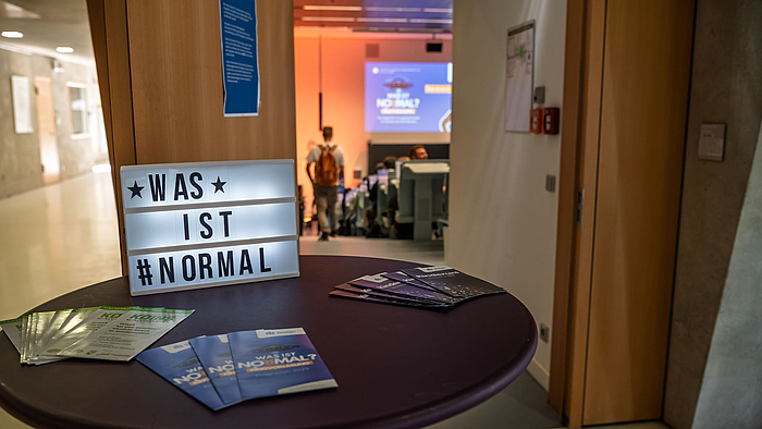 Auf einem runden Stehtisch vor einer geöffneten Hörsaaltür sind vor einem Leuchtschild mit dem Text „Was ist normal“ Flyer ausgebreitet. 