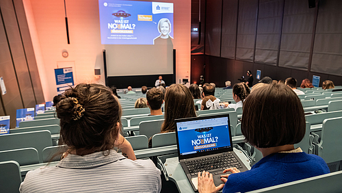 Sieben Mal stellt in diesem Sommersemester die Ringvorlesung die Frage „Was ist normal?“. Im fünften Vortrag gibt sie Antworten, Denkanstöße und Diskussionsstoff im Blick auf unser Verständnis von Arbeit und auf ihre vielfältigen Formen. Blick von hinten auf das Publikum in einem Hörsaal. Auf dem Laptop einer Besucherin und projiziert an der Rückwand des Hörsaals ist die Titelfolie zu sehen unter anderem mit dem Text „Was ist normal?' und dem Porträt der heutigen Referentin.