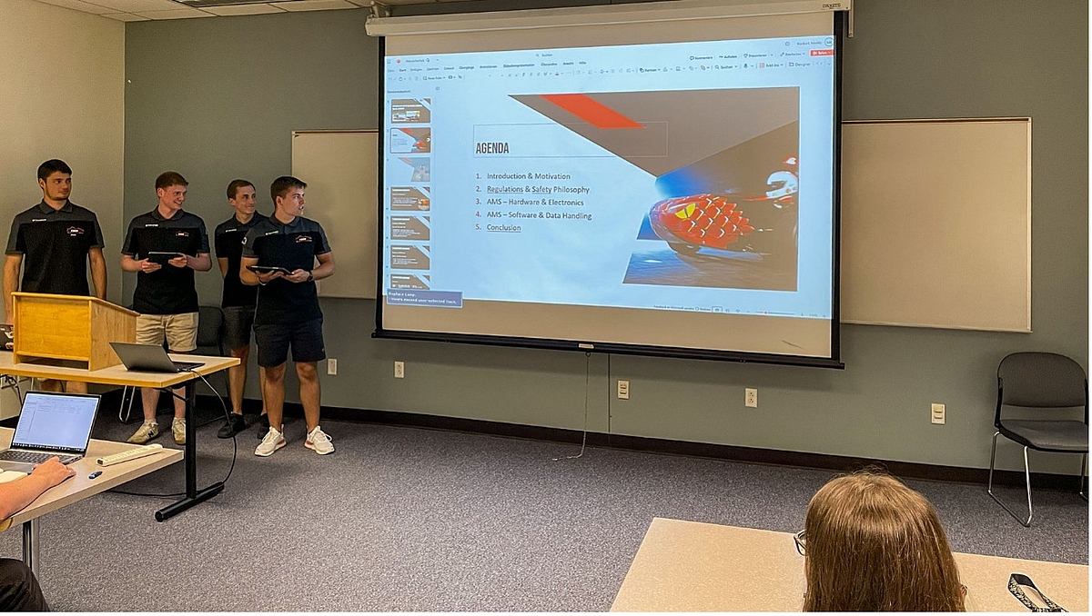 Mittweidaer Studierende präsentieren: hier zum Thema AMS – Application Management Services –bei der Entwicklung des TMM-Elektrorennwagens. Vier männliche Personen stehen neben einer Leinwand mit eine Powerpointpräsentation. Die Folie zeigt die Überschrift "Agenda" und zeigt das Vorderteil eines Rennwagens.