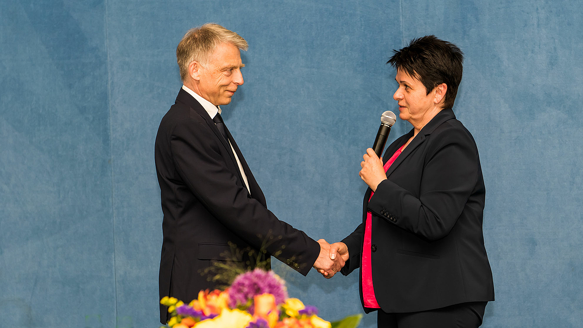 Das Foto zeigt zwei personen sich vor der blauen Rückwand der Sporthalle gegenüberstehend. Die weibliche Person rechts im Bild gratuliert der männlichen Person links mit Handschlag. Im Bildvordergrund sind unscharf bunte Blumen zu erkennen.