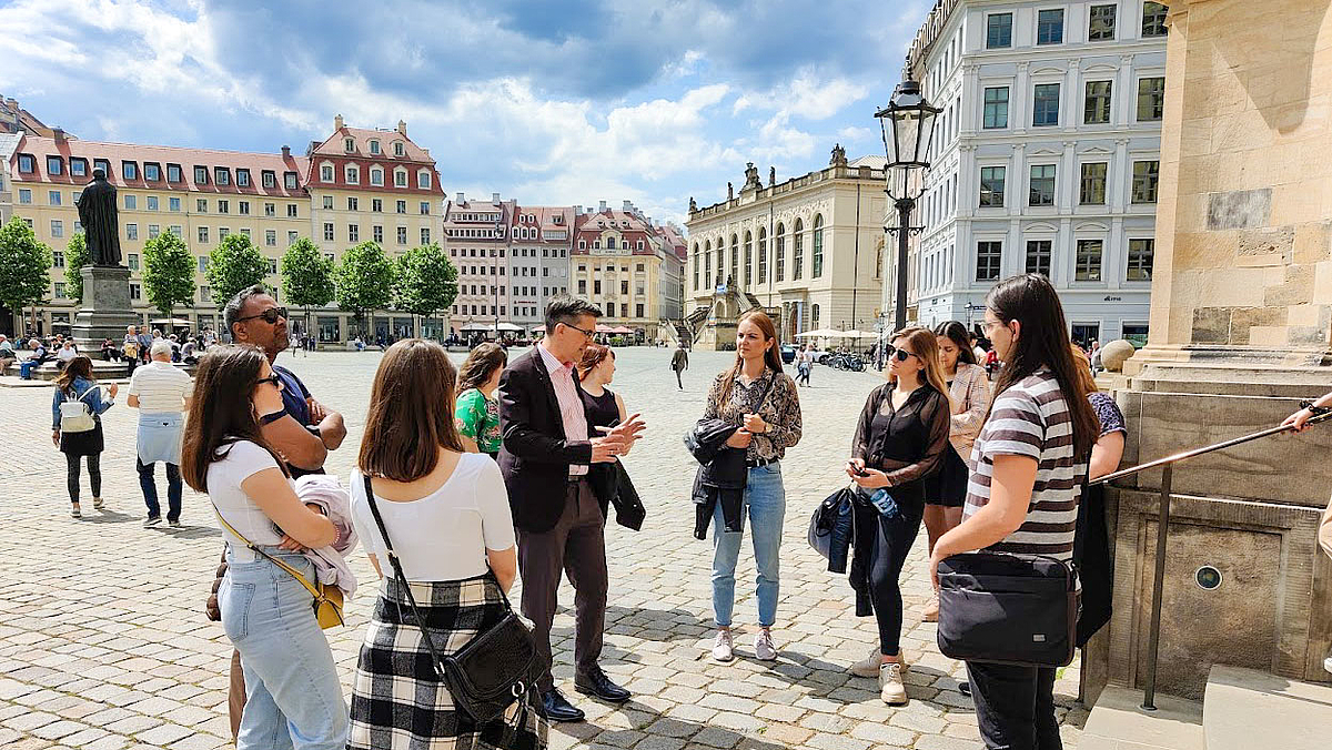 Das Foto zeigt eine Gruppe von 9 jungen Menschen auf dem Neumarkt in Dresden. Sie folgen den Erklärungen einer männlichen Person in ihrer Mitte.
