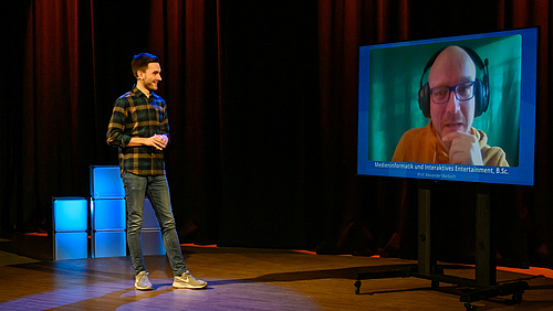 Live aus dem TV-Studio: Studienberater Maximilian Benda befragt Professor Alexander Marbach zum Studiengang "Medieninformatik und interaktives Entertainment". Das Foto zeigt einen jungen Mann und einen großen Monitor vor ienem dunklen Studiohintergrund. Der junge Mann wendet sich einer über den Monitor zugeschalteten Person zu.