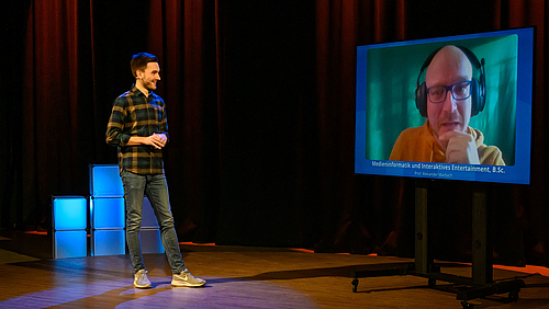Live aus dem TV-Studio: Studienberater Maximilian Benda befragt Professor Alexander Marbach zum Studiengang "Medieninformatik und interaktives Entertainment". Das Foto zeigt einen jungen Mann und einen großen Monitor vor ienem dunklen Studiohintergrund. Der junge Mann wendet sich einer über den Monitor zugeschalteten Person zu.