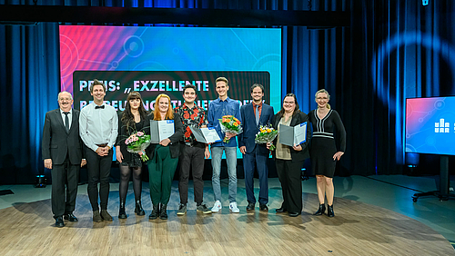 Große Bühne für großes Engagement beim Preis für exzellente Betreuung Studierender an der Hochschule Mittweida Gruppenfoto: Neun Personen stehen dicht nebeneinander auf einem niedrigen Podest in einem großen Studioraum. Drei von ihnen halten Blumensträuße und Urkunden in den Händen. Im Hintergrund ist vor einem in senkrechten Falten fallenden dunklem Vorhang ein sehr großes LED-Display, auf dem teilweise verdeckt von den Personen "Preis Exzellente Betreuung Studierender" zu lesen ist..