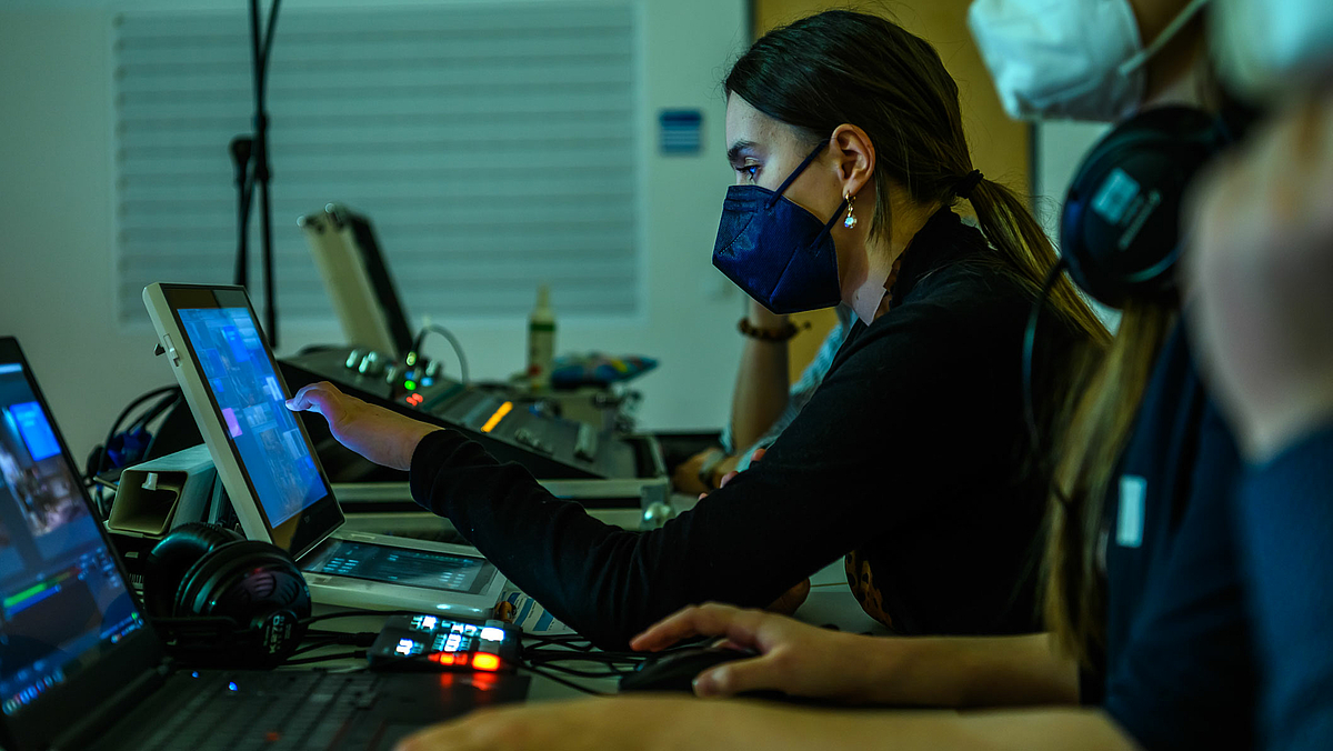 Das Foto zeigt von der Seite aufgenommen eine junge Frau mit FFP2-Maske vor einem mobilen Live-Videomischer. Im Vordergrund sind unscharf und angeschnitten ein Laptop sowie Hände und Kopf einer weiteren Person zu erkennen, im Hintergrund ein Tonmischpult.