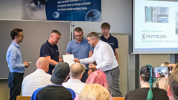 Preis für junge Gründer vom erfahrenen Gründer: Prof. Detlev Müller überreicht beim SAXEED-Gründerstammtisch die Auszeichnung an Hagen Grüttner (2.v.l.), Jan Bretschneider und Johannes Maus von ANTACON. Vor Publikum sind im Bildhintergrund fünf männliche Personen zu sehen. Eine von ihnen bekommt von einer anderen eine Urkunde überreicht (inhalt nicht erkennbar). Auf einem Monitor rechts neben der Personengruppe ist der Schriftzug ANTACON zu erkennen.