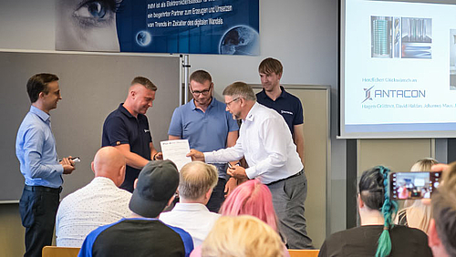 Preis für junge Gründer vom erfahrenen Gründer: Prof. Detlev Müller überreicht beim SAXEED-Gründerstammtisch die Auszeichnung an Hagen Grüttner (2.v.l.), Jan Bretschneider und Johannes Maus von ANTACON. Vor Publikum sind im Bildhintergrund fünf männliche Personen zu sehen. Eine von ihnen bekommt von einer anderen eine Urkunde überreicht (inhalt nicht erkennbar). Auf einem Monitor rechts neben der Personengruppe ist der Schriftzug ANTACON zu erkennen.