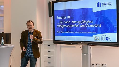 Impulsvortrag beim KI-Vernetzungstreffen: Der Mathematiker Professor Thomas Villmann von der Hochschule Mittweida brennt für smarte KI. Er und sein Institut sind Vorreiter in Sachsen. Das Foto zeigt eine männliche Person mit Mikrofon in der Hand links neben einem großen Display stehend. Dort ist eine Grafik zu sehen mit verschiedenen Logos, u.a. der Hochschule Mittweida und dem Text „Smarte KI … für hohe Leistungsfähigkeit, Interpretierbarkeit und Akzeptant. Prof. Thomas Villmann“.