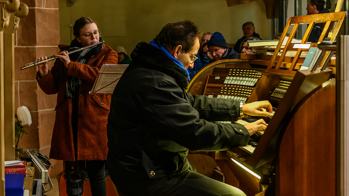 Links im Bild steht eine Musikerin mit Querflöte, rechts im Vordergrund Spieltisch mit ins Spiel vertieftem Organisten von der Seite.