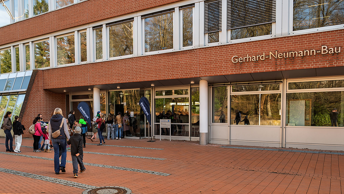 Menschen, darunter Kinder mit Rucksäcken, laufen zum Eingang des Gerhard-Neumann-Baus vor dem zwei Fahnen mit der Aufschrift „Kinderuni“ stehen.