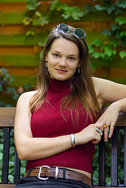 Eine junge Frau sitzt lässig auf einer Bank im Grünen, die Sonnenbrille ins Haar geschoben, und lächelt in die Kamera.