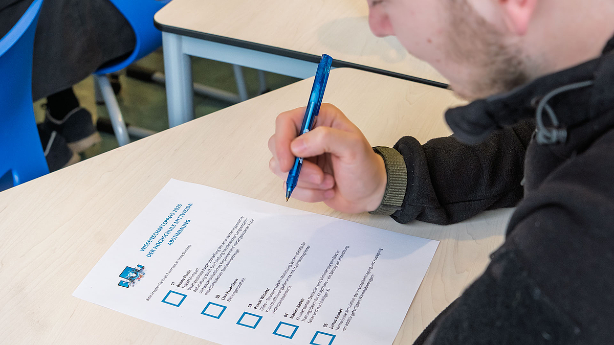 Das Publikum beim Tag der Forschung entscheidet mit. Blick auf eine männliche Person mit Kugelschreiber vor einem Stimmzettel mit fünf Wahlmöglickeiten.