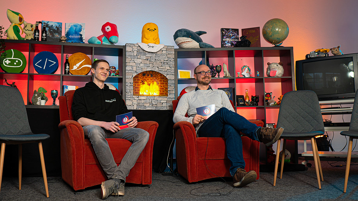Professor Alexander Marbach und Student Max Schlosser sitzen auf zwei roten Sesseln und lächeln in Richtung einer Kamera. Im Hintergrund steht ein Regal mit Game-bezogenen Plüschfiguren.