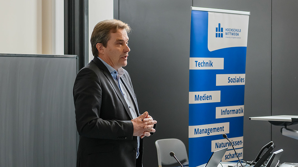 Eine männliche Person steht mit gefalteten Händen an einem Pult vor einer Tafel. Im Bildhintergrund steht ein Rollup mit dem Logo der Hochschule Mittweida und den Begriffen „Technik“, „Soziales“, „Medien“, Informatik“, Management“ und „Naturwissenschaft“.