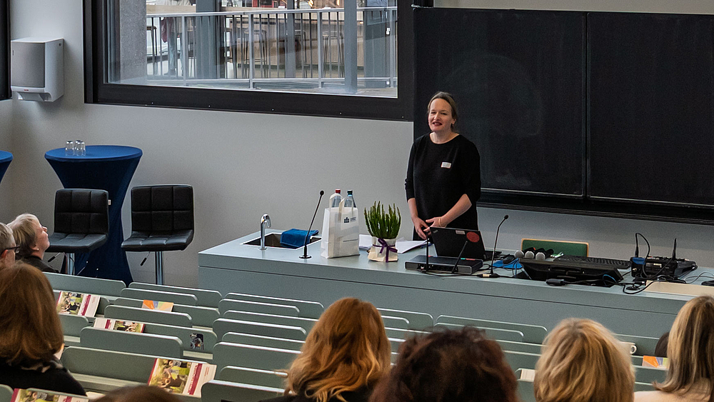 Weibliche Person lächelnd hinter einem Hörsaalpult. Im Bildvordergrund Köpfe von Personen von hinten in den ansteigenden Hörsaalbänken.