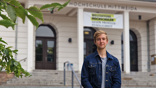 Nicholas Dean Olsen kam für ein Auslandssemester aus Shepherdstown in West Virginia, USA an die Hochschule Mittweida. Nicholas Dean Olsen steht vor dem Hauptgebäude der Hochschule Mittweida. Er hat blonde Haare und trägt eine blaue Jeans-Jacke.