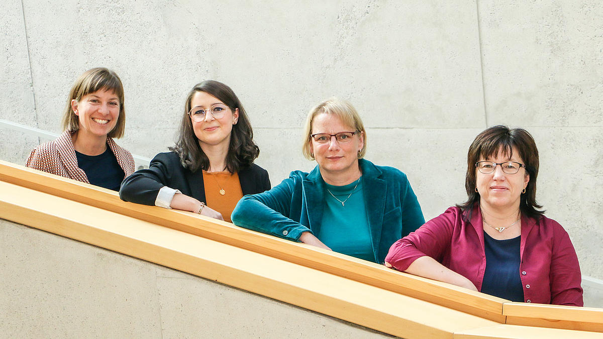 : Vier weibliche Personen stehen lächelnd auf einer nach links aufsteigenden Treppe.