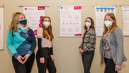 Patricia, Rosa Clara, Friederike und Juliane (v.l.) haben in ihrem Praxisprojekt eine Zufriedenheitsstudie für die Mitarbeitenden einer Werkstatt für behinderte Menschen erstellt. Das Foto zeigt vier Studentinnen Patricia, Rosa Clara, Friederike und Juliane vor einer Wand mit Forschungspostern.