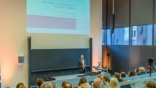 Seine Forschung geschieht auf einer abgeschirmten Insel hinter Stacheldraht, und eigentlich darf von dort nichts nach außen gelangen. In Mittweida aber berichtet Dr. Klaas Dietze von seiner Arbeit an Infektionskrankheiten. : Blick von seitlich hinten über die Köpfe der Zuschauenden auf den Dozenten, der vor der Tafel steht. Über ihm zeigt die Projektion die Titelfolie der Vorlesung: „One Health – Zeitenwende, wie wir Gesundheit verstehen? 5. Öffentliche Ringvorlesung. Hochschule Mittweida. Dr. Klaas Dietze.“