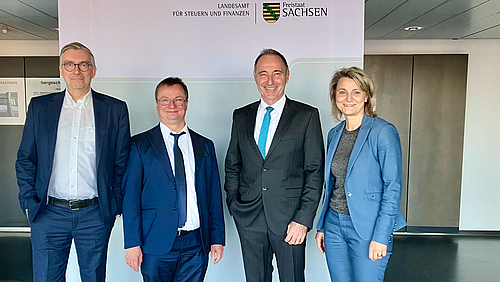 Arbeiten an der Forschung zum Einsatz von neuen Technolgien in der Finanzverwaltung zusammen: die Hochschule Mittweida und das Landesamt für Steuern und Finanzen Sachsen. (Foto: LFS) Drei männliche und eine weibliche Person in Anzügen stehen vor einem Rollup, auf dem zwichen dem sächsichen Wappen die Schriftzüge "Landesamt für Steuern und Finanzen" und "Freistaat Sachsen" zu lesen sind.
