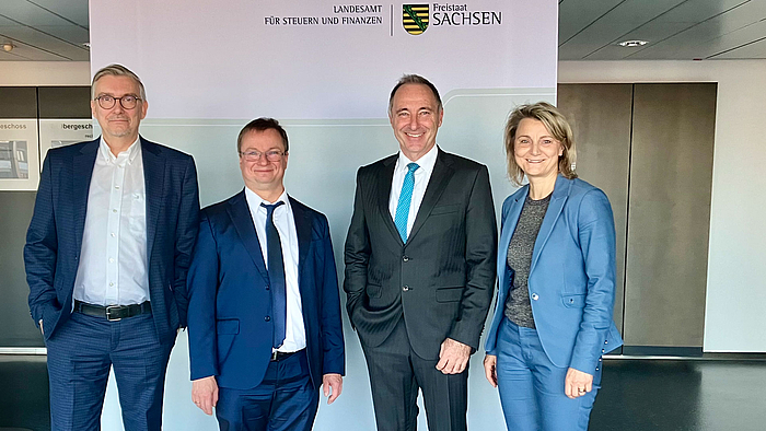 Arbeiten an der Forschung zum Einsatz von neuen Technolgien in der Finanzverwaltung zusammen: die Hochschule Mittweida und das Landesamt für Steuern und Finanzen Sachsen. (Foto: LFS) Drei männliche und eine weibliche Person in Anzügen stehen vor einem Rollup, auf dem zwichen dem sächsichen Wappen die Schriftzüge "Landesamt für Steuern und Finanzen" und "Freistaat Sachsen" zu lesen sind.