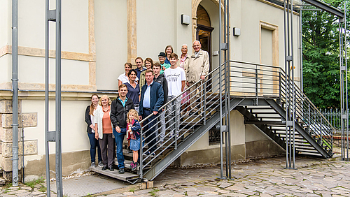 Familienbesuch mit drei Generationen: Carolin Zeller und Dr. Marion Stascheit (unten links) vom Hochschularchiv Mittweida begrüßen Nachkommen von Alfred Udo Holzt, dem Direktor des Technikum Mittweida von 1892 bis 1936. Das Foto zeigt 15 Personen unterschiedlichen Alters auf der Außentreppe der Direktorenvilla der Hochschule Mittweida. Neben den beiden Mitarbeiterinnen des Hochschularchivs Carolin Zeller und Dr. Marion Stascheit handelt es sich um Mitglieder der Familie Knieper, Nachkommen des zweiten Direktors des Technikum Mittweida Alfred Udo Holzt (1859–1945)