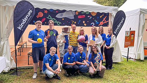 Hochschule Mittweida auf (dritter) Mission. Das Team für Experimentieren im Park macht Kinder neugierig auf Naturwissenschaft. 12 Personen unterschiedleichen geschlechst und Alters steh oder knien vor ienem offenen weißen Pavilion. Einige targen weiße Laborkittel, eine person hat inen Imkerhut auf. Links und rechts von der Gruppe stehen zwei Teardrop-Flags mit der Aufschrift "kinderuni".
