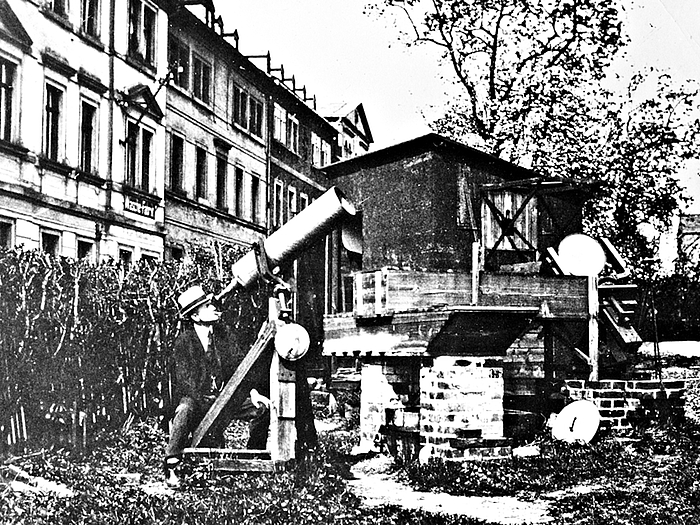 Bernhardt Schmidts schaut an seiner Sternwarte in Mittweida durch ein Teleskop. Er sitzt auf einer Wiese, im Hintergrund ist ein Bretterschuppen zu sehen, links einige mehrgeschossige Häuser.