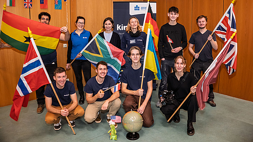 Zehnhn Personen stehen und knien im Hörsaal, einige halten verschiedene Nationalflaggen hoch und lächeln. Hinter der Gruppe ist ein Kinderuni-Rollup.