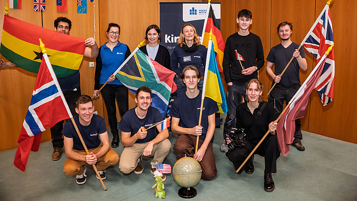 Mission completed: Das Kinderuni-Team der Hochschule macht Globalisierung und andere spannende Themen erlebbar. Zehnhn Personen stehen und knien im Hörsaal, einige halten verschiedene Nationalflaggen hoch und lächeln. Hinter der Gruppe ist ein Kinderuni-Rollup.