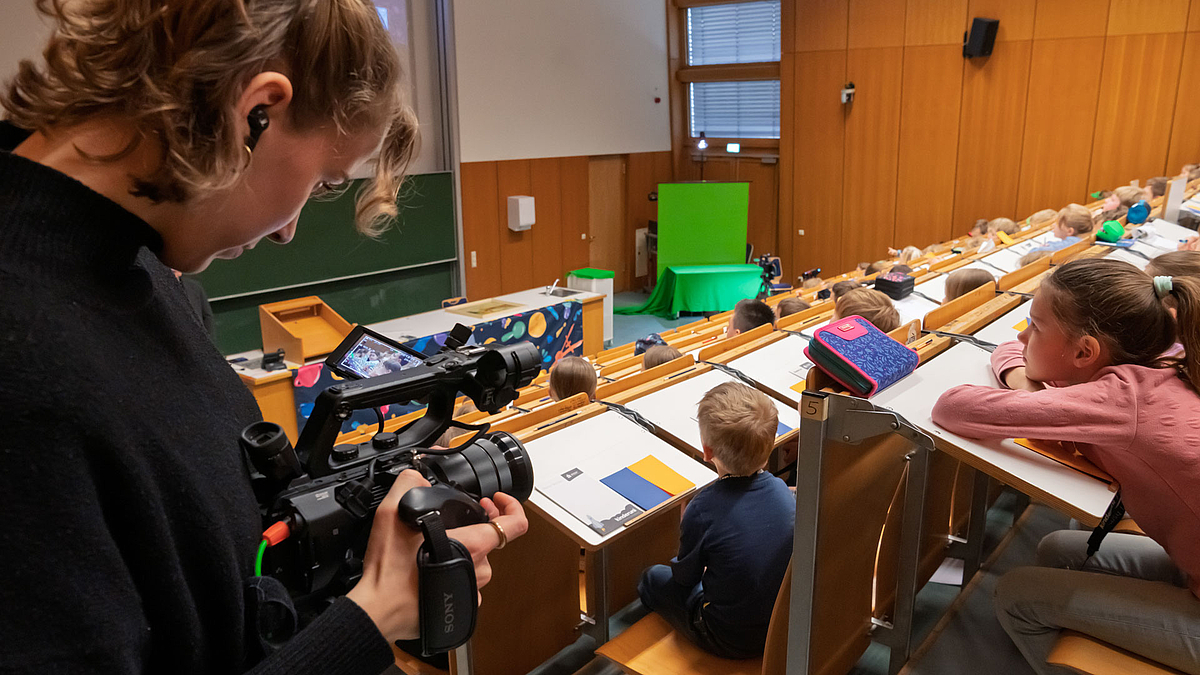 Eine Frau mit einer Kamera filmt Kinder in einem Hörsaal. Im Hintergrund sieht man Kinder, die aufmerksam nach vorne schauen.