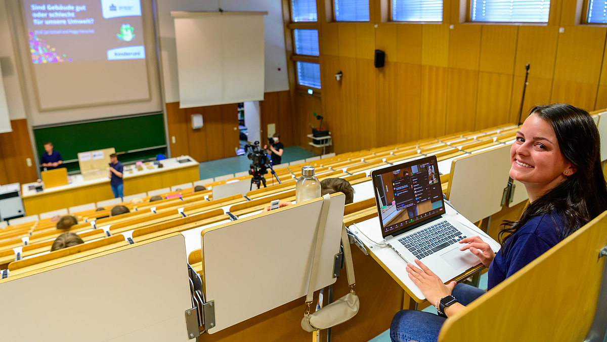 Das Foto zeigt eine junge Frau vor einem Laptop in der obersten Bankreihe des Hörsaals, fotografiert von hinten über die Schulter. Sie wendet den Blick zur Kamera und lächelt. Im Bildhintergrund sind die absteigenden Bankreihen des Hörsaals zu erkennen.