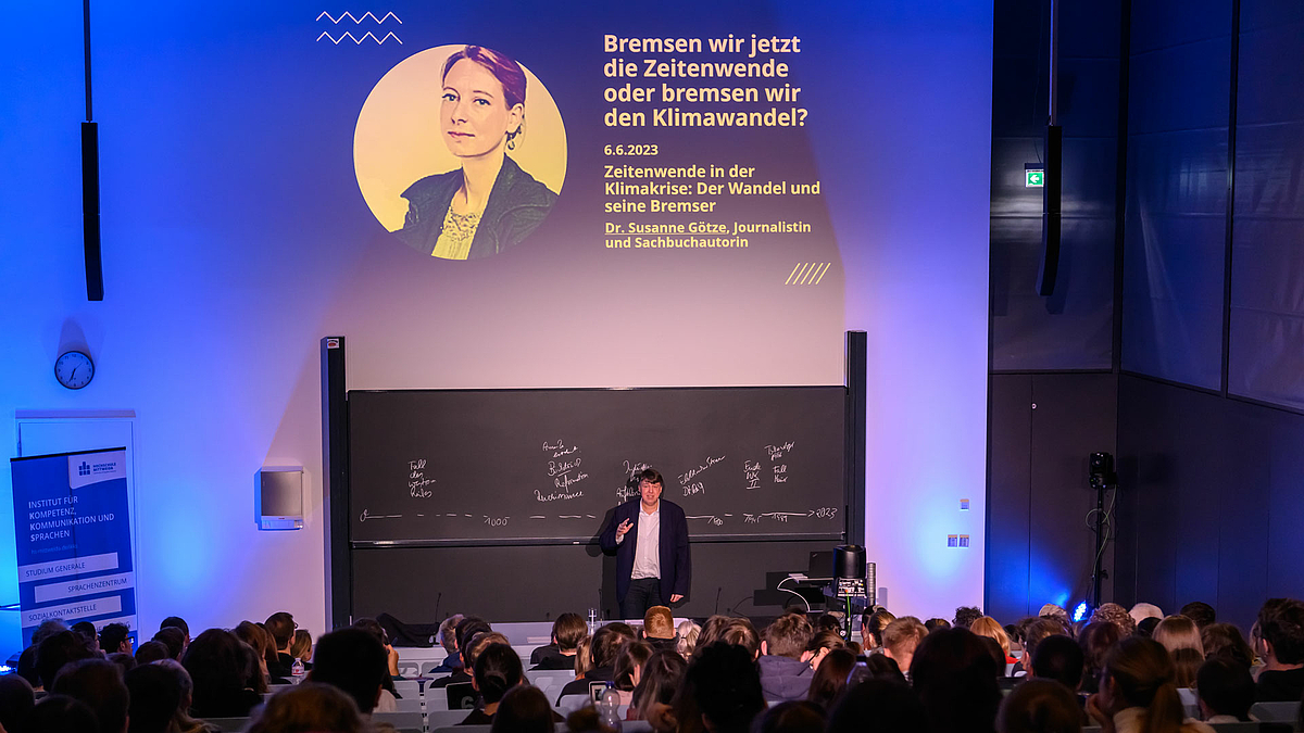 Blick über die Köpfe der Zuhörenden im Hörsaal hinweg nach vorn auf den Dozenten hinter dem Pult. Über ihm ist eine Folie projiziert mit dem Foto einer weiblichen Person links und rechts dem Text: „Bremsen wir jetzt die Zeitenwende oder bremsen wir den Klimawandel? 6.6.2023. Zeitenwende in der Klimakrise: Der Wandel und seine Bremser. Dr. Susanne Götze, Journalistin und Sachbuchautorin.“