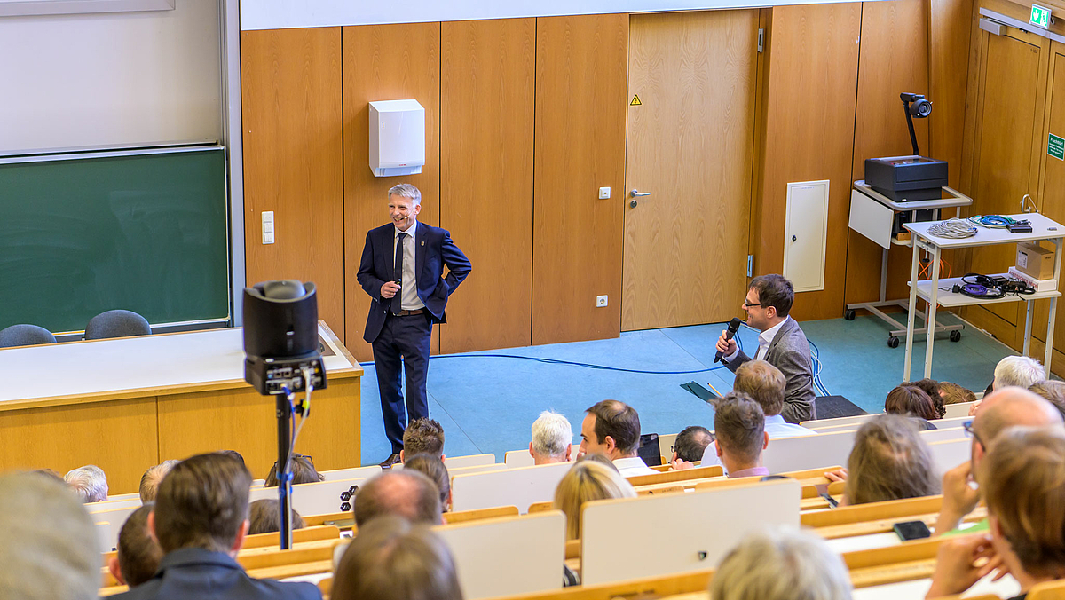 Das Foto blickt von oben in den  vollbesetzten Hörsaal und zeigt am unteren Ende des Hörsaals eine männliche Person im blauen Anzug und Krawatte lachend dem Publikum zugewandt. Rechts im Bild ist ein männliche Person stehend in den Bankreihen mit einem Mikro in der Hand, die offensichtlich eine Frage gestellt hat.