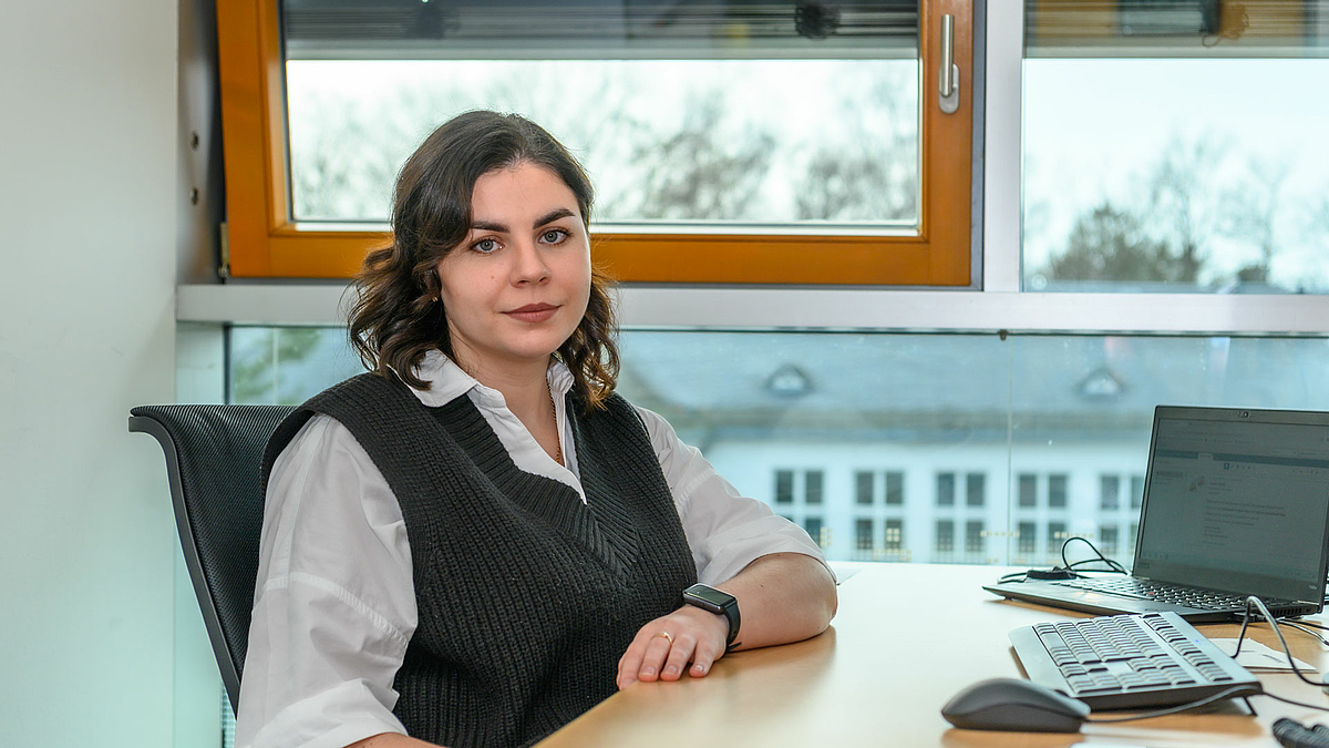 Daryna Karpiuk sitzt an einem Schreibtisch mit Laptop vor einer Fensterfront. Sie trägt Bluse und Wollweste und blickt freundlich, aber bestimmt in die Kamera.